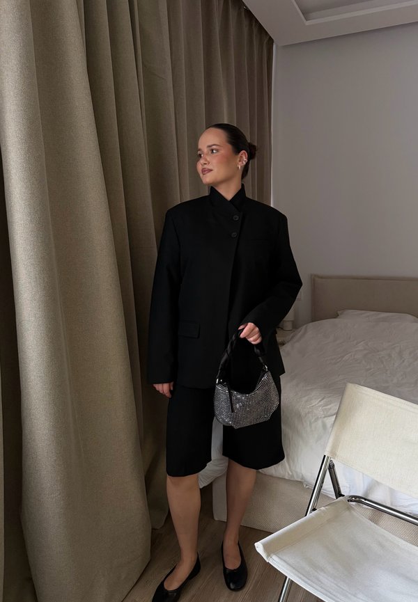 Mujer con blazer negro y pantalones cortos sosteniendo un pequeño bolso, de pie cerca de unas cortinas beige en un dormitorio con cama blanca y silla.