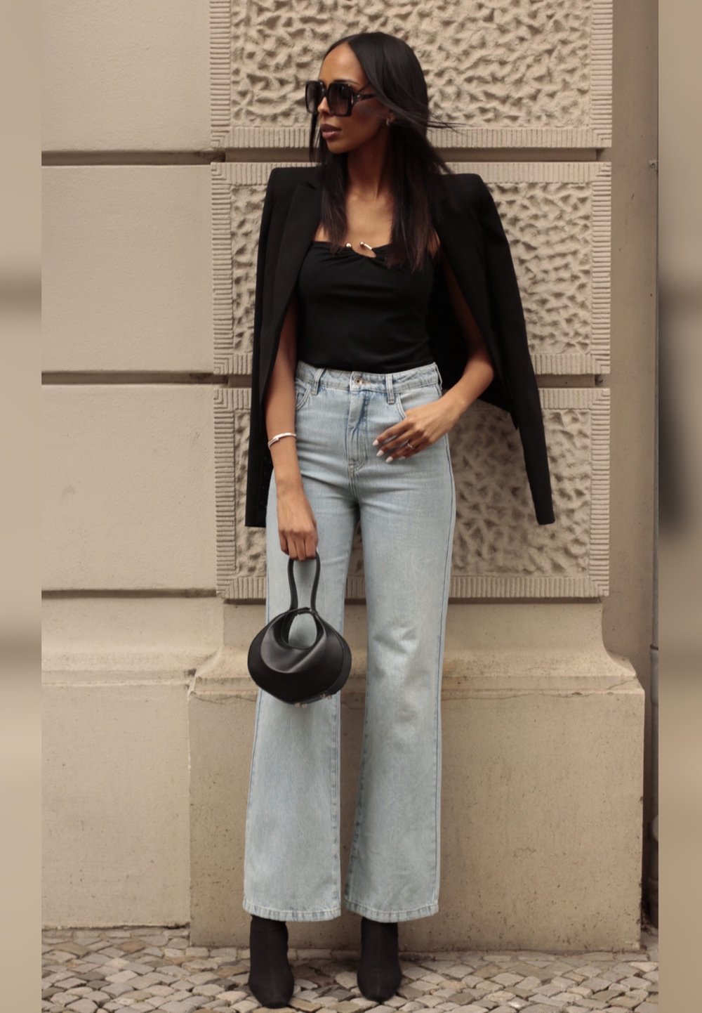 Črn blejzer, prilegajoča črna majica, svetlo modri široki kavbojki, črne čevlje s peto in okrogla črna torbica. Ozadje z teksturiranim vzorcem.