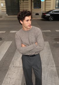 Jauns vīrietis ar cirtainiem matiem, ģērbies pelēkā džemperī un tumšās biksēs, stāv pilsētas pārejā ar saliktām rokām, skatoties pa labi.