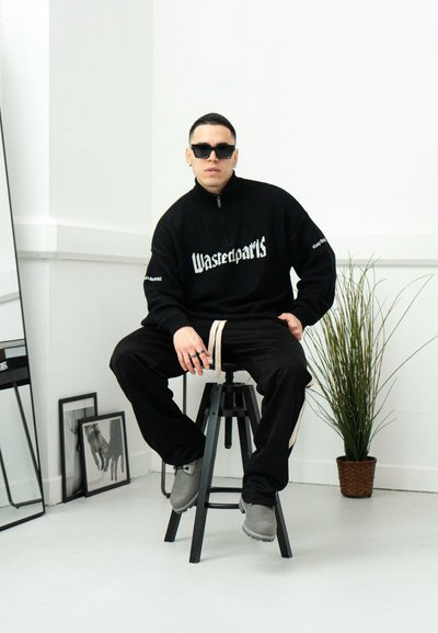 Sudadera negra con cremallera y el logo "Wastedparts", combinada con pantalones negros y zapatos grises. Las características incluyen un ajuste relajado, mangas largas y una textura gruesa.