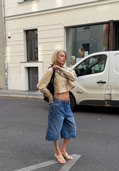 Chaqueta beige recortada con cierre de cremallera, pantalones cortos de mezclilla de pierna ancha azul y sandalias beige. Mochila negra llevada en un hombro. Entorno urbano.