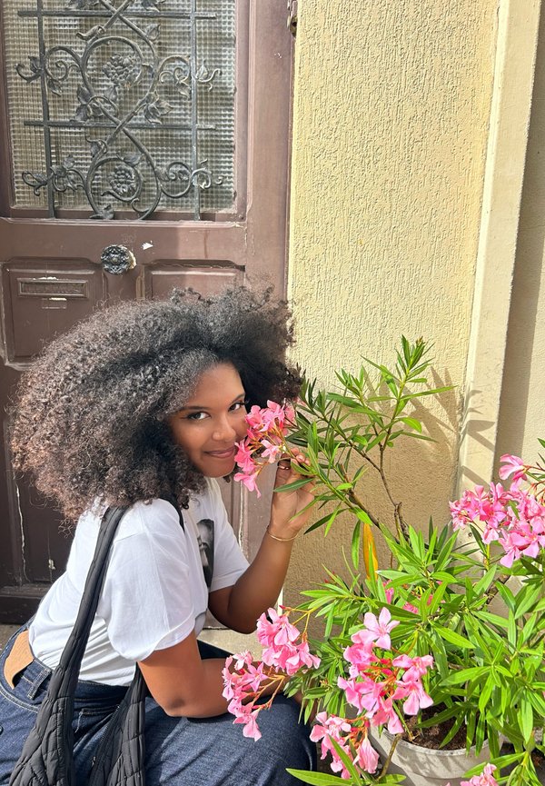 Kvinde med krøllet sort hår læner sig mod rosa oleanderblomster, iført en hvid T-shirt og en sort taske, ved en brun trædør med metalbisser.