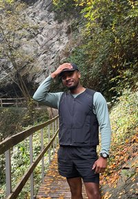 Idrettsantrekk som inkluderer en svart vest over en teal langermet skjorte, kombinert med svarte shorts. Solid gangvei omgitt av grøntområder.