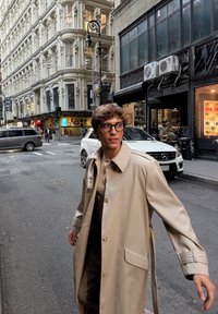 Bežový kabát po kolená s veľkými gombíkmi a vreckom, nosený cez hnedý outfit, na pozadí mestského ulice.