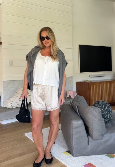 Mujer con blusa blanca, pantalones cortos de seda, suéter gris sobre los hombros, bailarinas negras, gafas de sol y sostiene un bolso negro en una sala de estar moderna.