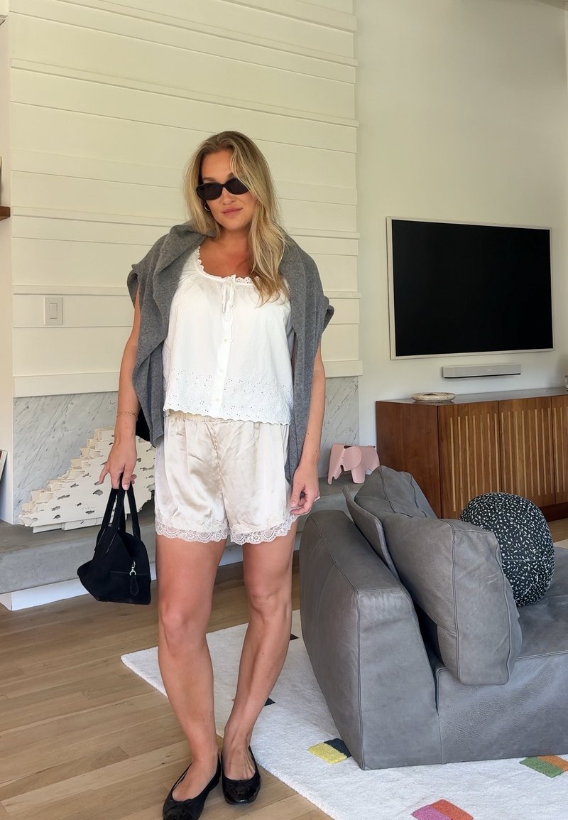 Γυναίκα φοράει λευκό πουκάμισο, μεταξένιες βερμούδες, γκρι πουλόβερ περασμένο στους ώμους, μαύρα φλατ παπούτσια, γυαλιά ηλίου και κρατά μια μαύρη τσάντα σε ένα σύγχρονο σαλόνι.