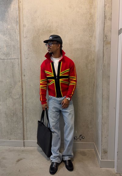 Hombre con suéter rojo con cremallera y patrón geométrico amarillo y negro, jeans claros, gorra negra, gafas, y sosteniendo un bolso negro de tela, de pie en el interior.