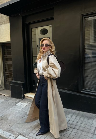 Mujer con cabello largo y rubio, usando gafas de sol, abrigo beige, blusa blanca, vaqueros oscuros y llevando una mochila negra, de pie en una acera de la ciudad.