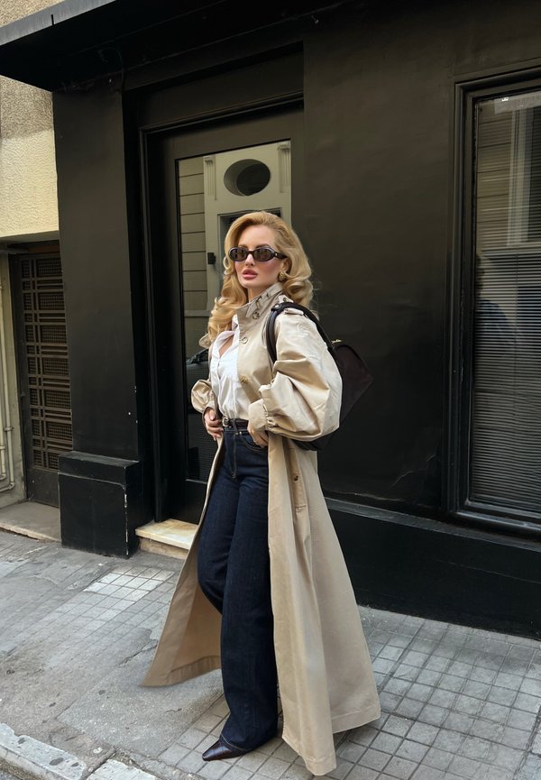 Žena s dlhými blond vlasmi, ktorá nosí slnečné okuliare, béžový trenčkot, bielu blúzku, tmavé rifle a nesie čierny batoh, stojí na mestskom chodníku.