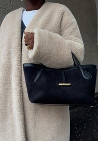 Άτομο που φοράει μπεζ παχύ παλτό, κρατώντας μια μαύρη τσάντα από σουέντ με δερμάτινες λαβές και χρυσές λεπτομέρειες, φορώντας πολλά δαχτυλίδια στο αριστερό χέρι.