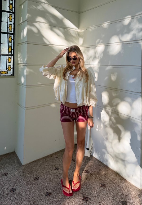 Young woman in sunglasses, white jacket, and striped shorts stands indoors near a stained glass window with dappled sunlight on walls.
