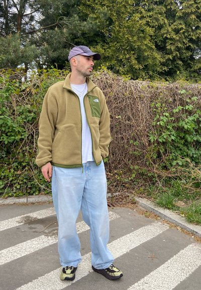 Hombre con gorra, chaqueta de forro polar verde, camisa blanca y vaqueros azul claro está en un paso de cebra junto a un seto frondoso.