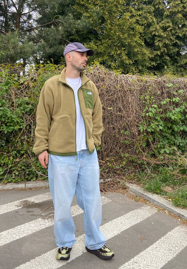 Muž s čepicí, zelenou fleecovou bundou, bílou košilí a světle modrými džínami stojí na přechodu u listnatého živého plotu.