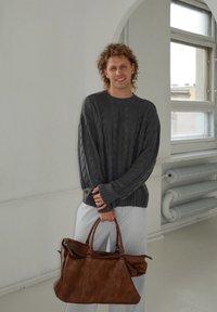 Suéter cinza com padrão de malha, calças brancas com riscas e uma mala de couro castanho com duas alças. Interior brilhante com luz natural.