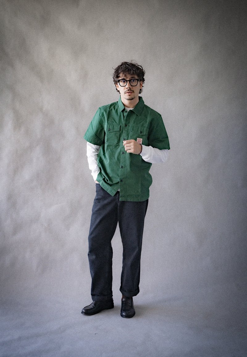 Verde camicia a maniche corte con due tasche sul petto e un'etichetta, indossata sopra a una camicia bianca a maniche lunghe; abbinata a pantaloni scuri e scarpe nere.