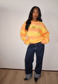 Sweatshirt dégradé orange et jaune avec un logo circulaire, accompagné d'un jean large bleu foncé aux ourlets retroussés et de talons noirs pointus.