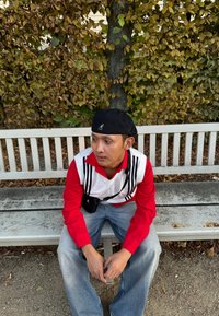 Κόκκινο μακρυμάνικο πουκάμισο με λευκό γιακά και μαύρες ρίγες, σε συνδυασμό με φαρδιά μπλε τζιν. Φοράει μαύρη μπερές και μικρή μαύρη τσάντα. Καθισμένος σε ένα παγκάκι.