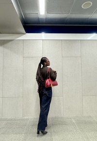 Haut noir à manches longues, jeans bleu ample et sac à main rouge. Cheveux tressés, debout contre un mur gris avec un sol carrelé.