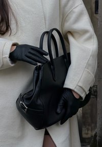 Musta nahkainen käsilaukku, jossa on kaksi kahvaa, sileä pinta ja vetoketjullinen suljenta. Yhdistetty mustiin hansikkaisiin ja vaaleanväriseen takkiin.