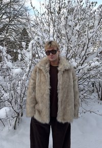 Manteau en fausse fourrure beige clair, avec une texture duveteuse et un large col, porté sur un haut foncé et assorti à un pantalon ample foncé. Fond enneigé.