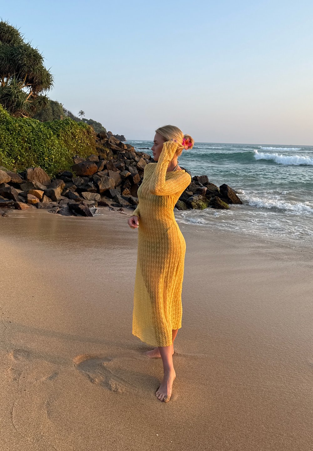 Femme en longue robe jaune, pieds nus, debout sur une plage de sable près des rochers, avec des vagues océaniques et de la verdure en arrière-plan au coucher du soleil.