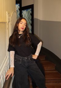 Zwart t-shirt, lange mouwen van kant, zwarte high-waist jeans, zilveren ketting, staand bij een trap, met decoratief glas-in-lood achter.