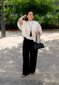 Mujer de pie al aire libre con pantalones anchos negros, blusa blanca suelta, bolso negro y collar de flecos largos, sosteniendo gafas de sol en la mano.