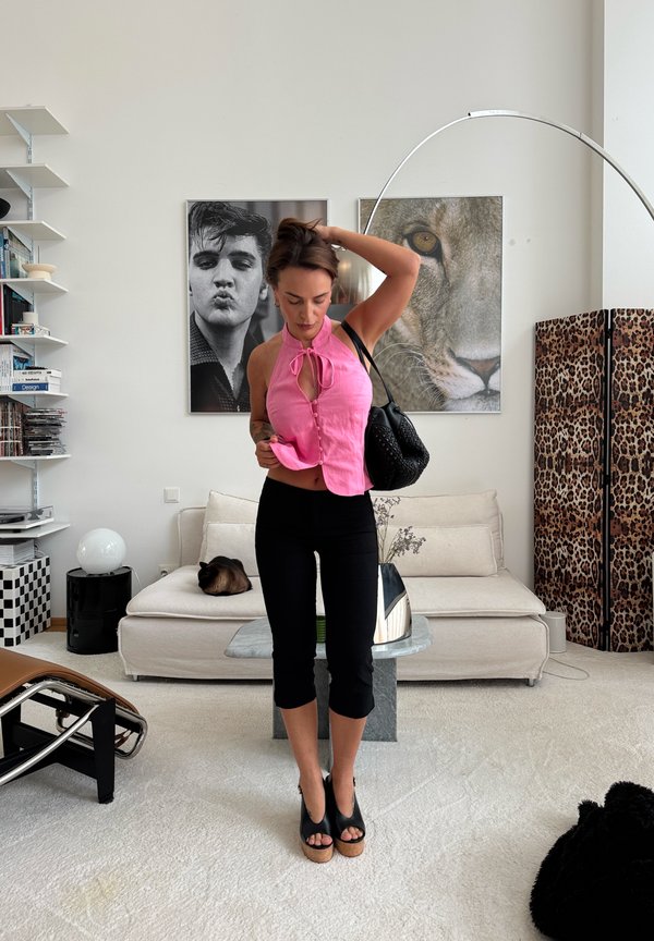 Kvinde i lyserød ærmeløs top og sorte bukser retter sit hår i en stilfuld stue med kat på sofaen, store portrætter af en løve og en mand samt en skærm med leopardprint.