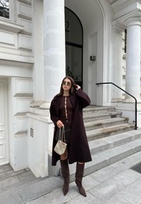 Palton lung de culoare vin roșu, bluză cu textură ribbed și butoane, fustă mini, cizme lungi din suede maro. Geantă transparentă cu design texturat. Fundalul prezintă scări și coloane.
