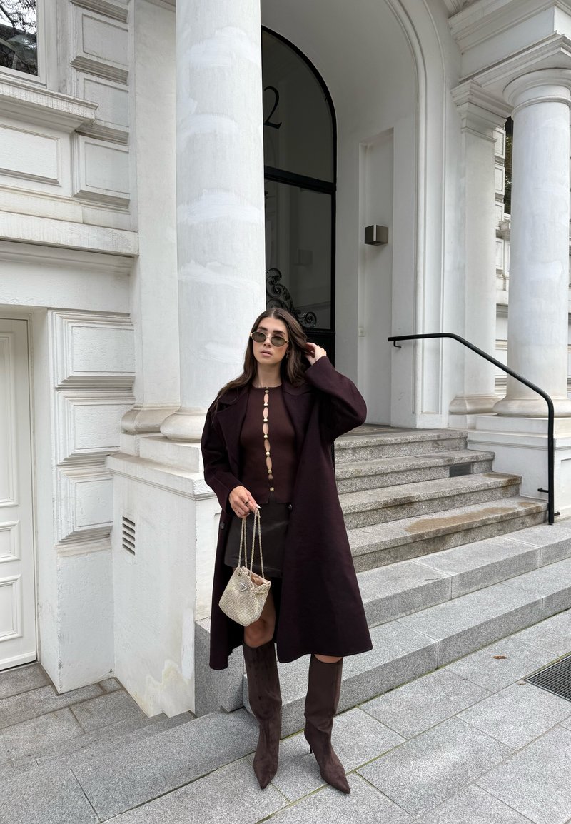 Abrigo largo burdeos, top acanalado con botones, falda corta, botas altas de ante marrón. Bolso transparente con diseño texturizado. El fondo presenta escalones y columnas.