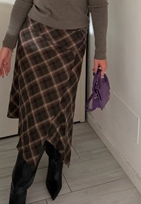 Brązowa spódnica w kratę wiązana, z asymetrycznym dołem, zestawiona z czarnymi botkami na szpic i małą fioletową torebką.