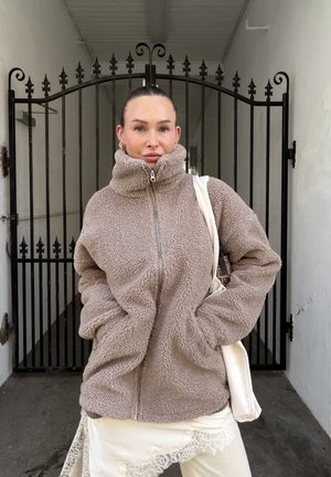 Mujer con el cabello recogido que lleva una chaqueta de forro polar beige cerrada con cremallera, falda blanca con encaje y lleva un bolso tote blanco, de pie frente a una puerta de hierro.