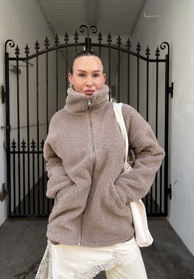 Mujer con el cabello recogido que lleva una chaqueta de forro polar beige cerrada con cremallera, falda blanca con encaje y lleva un bolso tote blanco, de pie frente a una puerta de hierro.