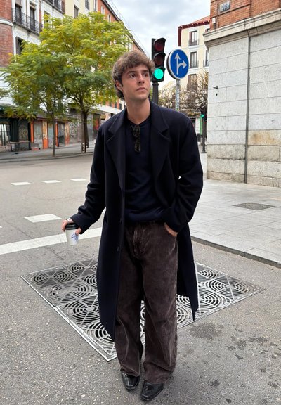 Abrigo largo negro sobre un suéter oscuro, combinado con pantalones anchos de pana marrón y zapatos negros puntiagudos. Sostiene un vaso de café desechable.