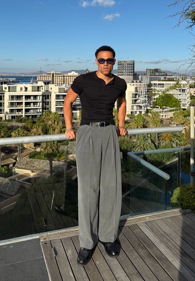 Hombre vestido con polo negro y pantalones anchos grises apoyado en una barandilla, con un paisaje urbano y un cielo azul despejado de fondo.