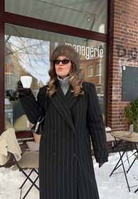 Femme portant un manteau à fines rayures, un chapeau en fourrure et des lunettes de soleil, tenant une tasse blanche devant un café avec de la neige au sol.