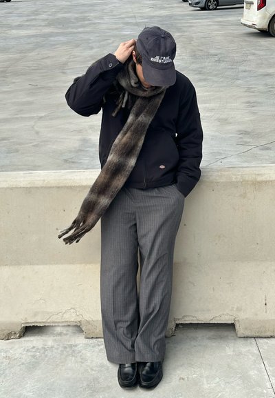 Chaqueta negra, pantalones de rayas grises, bufanda oscura y zapatos negros. Una gorra con texto completa el atuendo. De pie contra una barrera de concreto.