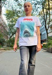 Weißes T-Shirt mit Grafik eines Haifischs und dem Schriftzug "JAWS", kombiniert mit grauen Jeans. Lässiges Design in einer natürlichen Umgebung im Freien.