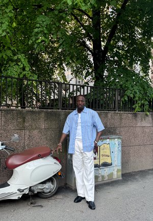 Un hombre con una camisa de rayas azul claro y mangas cortas, y pantalones blancos, está junto a un scooter blanco con un asiento marrón, frente a una pared texturizada.