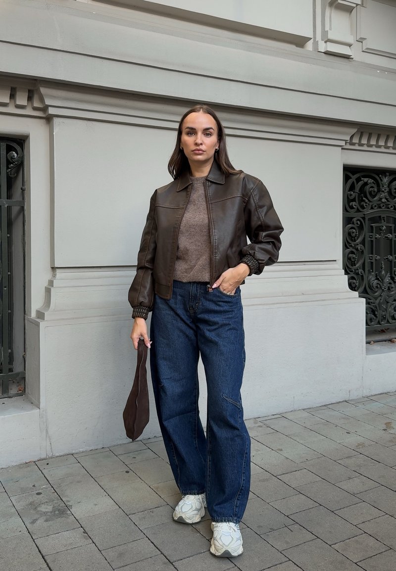 Femme debout sur le trottoir portant une veste en cuir marron, un pull marron, un jean bleu à jambes larges, des baskets blanches, tenant un sac marron dans une main.