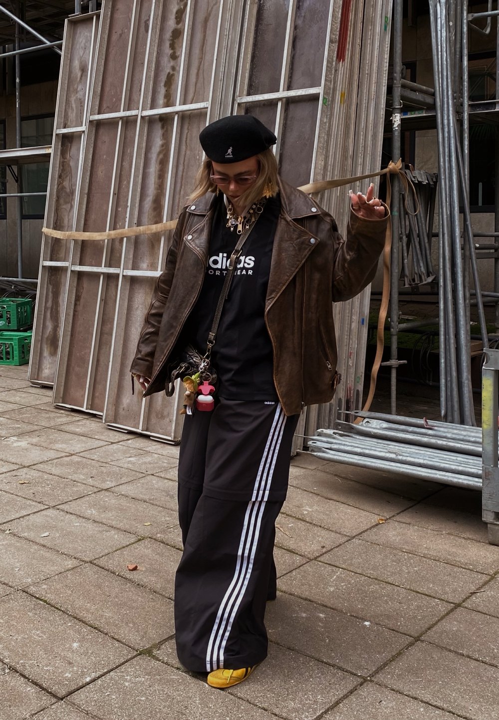 Barna bőrkabát, fekete Adidas póló, fekete széles nadrág fehér csíkokkal, sárga lábbeli, valamint kiegészítők, beleértve színes kulcstartókat.