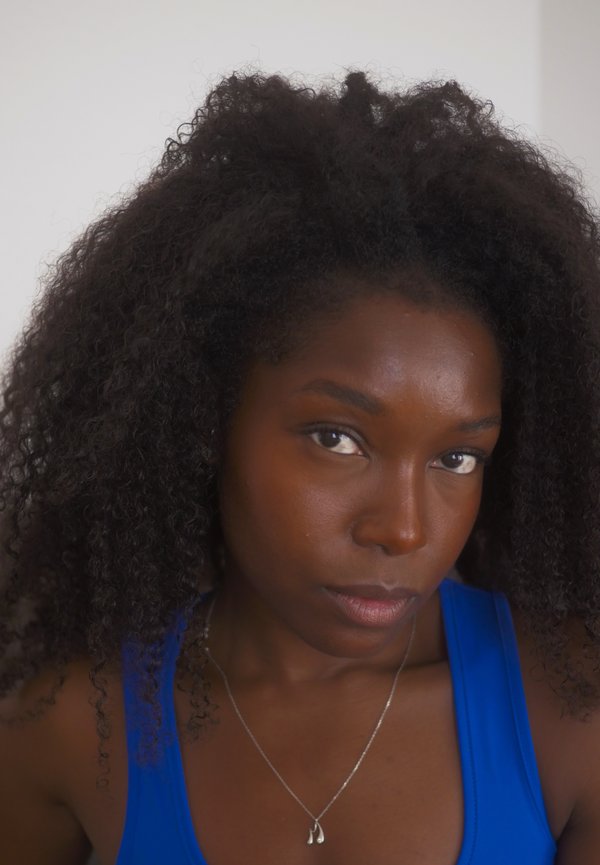 Femme aux cheveux noirs bouclés portant un haut bleu et un collier argenté, regardant directement la caméra avec une expression neutre sur un fond uni.