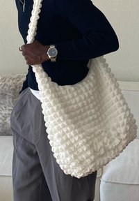 Osoba ubrana w granatowy sweter i szare spodnie, trzymająca dużą białą teksturowaną torbę na ramię w pomieszczeniu.