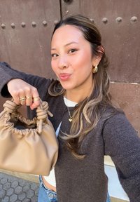 Beżowa torebka z marszczonym górnym brzegiem i zawiązanymi uchwytami. Nosi ją osoba w ciemnym swetrze i jasnej bluzce, na tle o fakturowanej powierzchni.