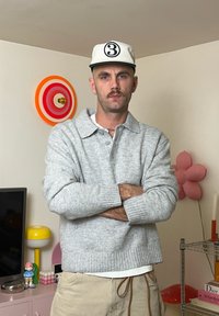 Helehall halli kampsun, millel on kraega disain, nööbidetail ja soonikkoes varrukasäär. Koos beezide pükste ja valge nokamütsiga, millel on must logo.