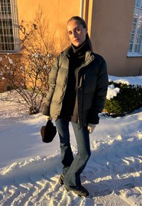 Puffjacke in Dunkelgrün, schwarzer Rollkragenpullover, blaue ausgestellte Jeans, schwarze Handschuhe, schwarze Tasche; verschneiter Boden und frostiger Hintergrund.