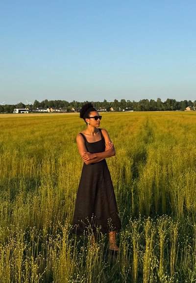Vestido midi negro de lino con tirantes de espagueti, que presenta una falda acampanada. La modelo está de pie en un campo de hierba alta amarilla-verde bajo un cielo despejado.