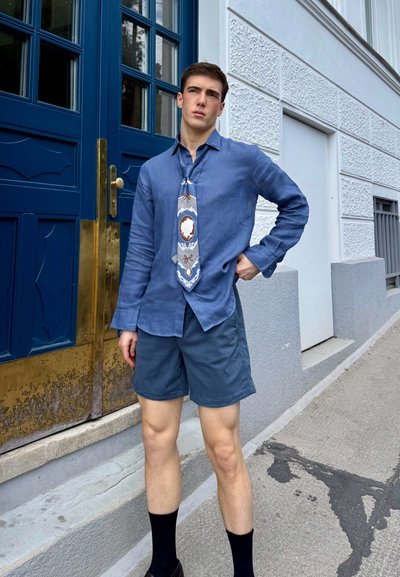 Camisa de lino azul claro, corbata con patrón en gris y blanco, pantalones cortos oscuros y calcetas negras. De pie junto a una puerta de entrada azul con herrajes dorados.