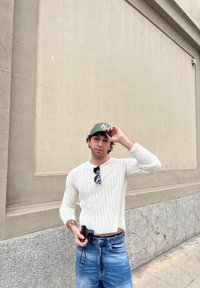 Hvid ribbet sweater med lange ærmer, lyseblå slidte jeans, holder et lille sort kamera og bærer en grøn kasket.