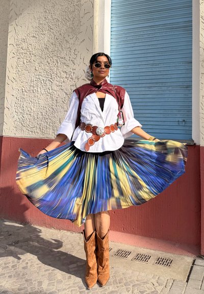 Mujer con gafas de sol, blusa blanca, chaleco de cuero, cinturón ancho marrón, falda plisada colorida y botas marrones, de pie junto a una pared texturizada y una persiana azul.